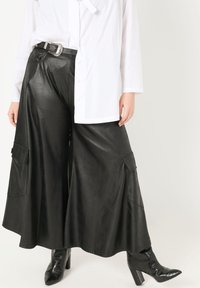 Schwarze, weit geschnittene Hose aus Kunstleder mit zwei seitlichen Taschen, kombiniert mit einem weißen Hemd mit Knöpfen. Schwarze strukturierte Knöchelschuhe.