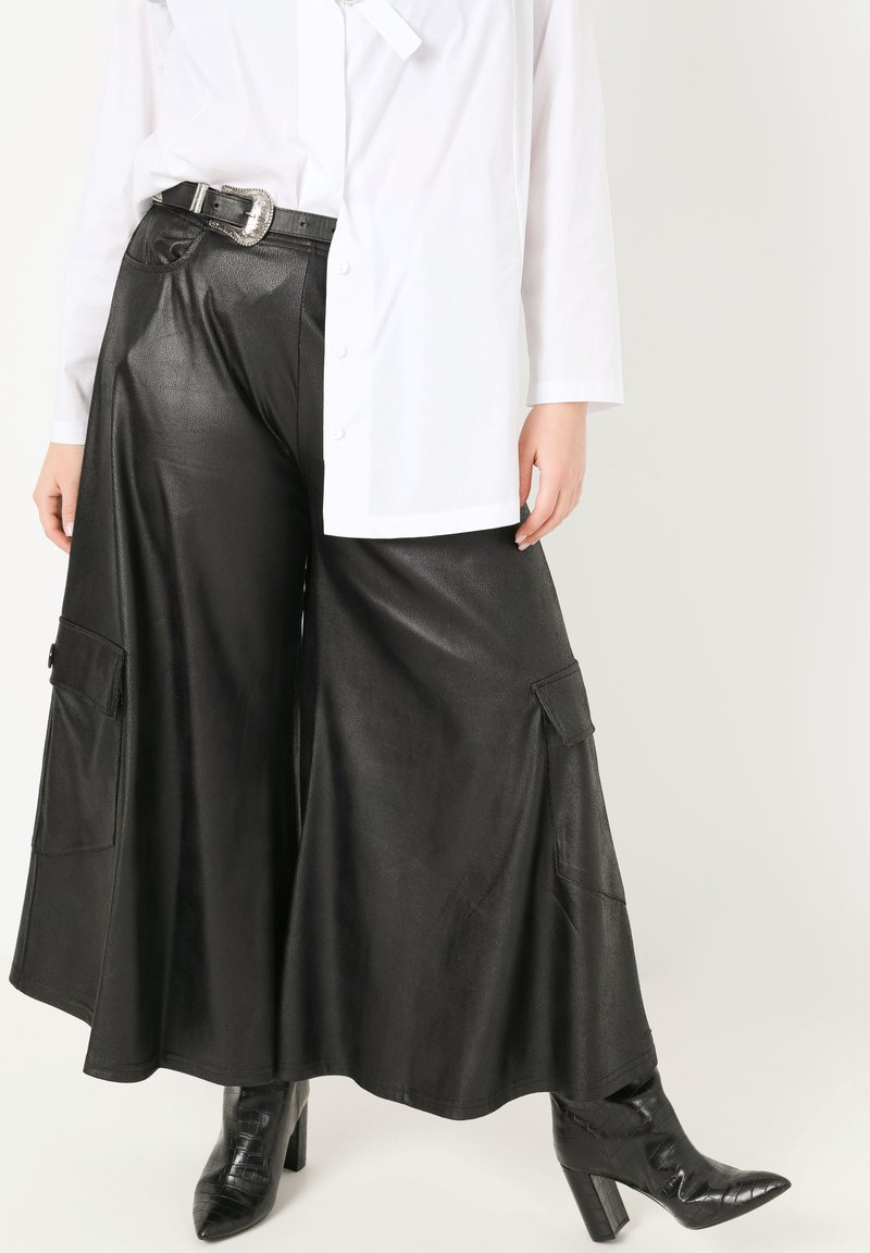 Schwarze, weit geschnittene Hose aus Kunstleder mit zwei seitlichen Taschen, kombiniert mit einem weißen Hemd mit Knöpfen. Schwarze strukturierte Knöchelschuhe.