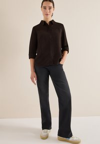 Schwarze Knopf-up-Hemd, lange Ärmel hochgerollt, kombiniert mit dunkelgrauen Jeans und weißen Sneakers mit beigen Akzenten. Einfaches, lässiges Outfit.