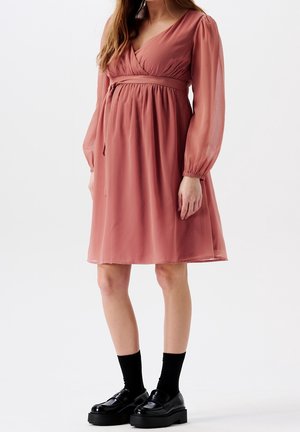 Femme portant une robe rose poudrée mi-longue à manches longues transparentes, des chaussettes noires et des mocassins compensés noirs sur fond blanc.