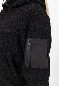 Haut à capuche en polaire noir avec une surface texturée et une poche zippée sur la manche gauche, doté d'une capuche réglable et de détails cousus.