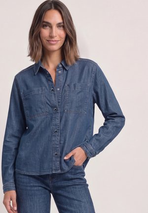 Frau, die ein blaues Jeanshemd mit Knöpfen und blaue Jeans trägt, steht mit der rechten Hand in der Tasche vor einem schlichten hellen Hintergrund.