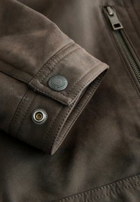Ärmel einer braunen Lederjacke mit Druckknopf und Reißverschluss, aus strukturiertem Stoff und mit kontrastierenden Nähdetails.