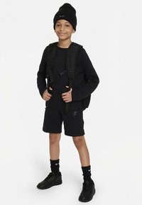 Melns Nike komplekts ar garām piedurknēm un šortiem, melna cepure un sporta apavi. Aprīkots ar melnu mugursomu.