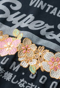 Tessuto nero con testo grigio presenta ricami floreali in rosa, beige e verde con dettagli di cucitura e un contorno arancione.