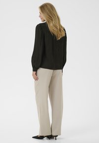 Frau mit langen blonden Haaren, die eine schwarze Bluse mit langen Ärmeln, bequeme beigen Hosen und schwarze flache Schuhe trägt, steht mit dem Rücken zu uns.