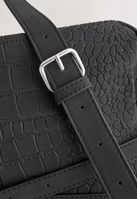 Melnas mākslīgās ādas soma ar krokodila reljefa tekstūru, regulējamu siksnu ar sudraba sprādzi un izsmalcinātu šuvju izstrādi.