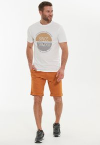 Weiße T-Shirt mit Grafikdesign, kombiniert mit orangefarbenen Shorts mit umgeschlagenem Saum. Die Schuhe haben graue und schwarze Akzente. Lässiges Sommeroutfit.