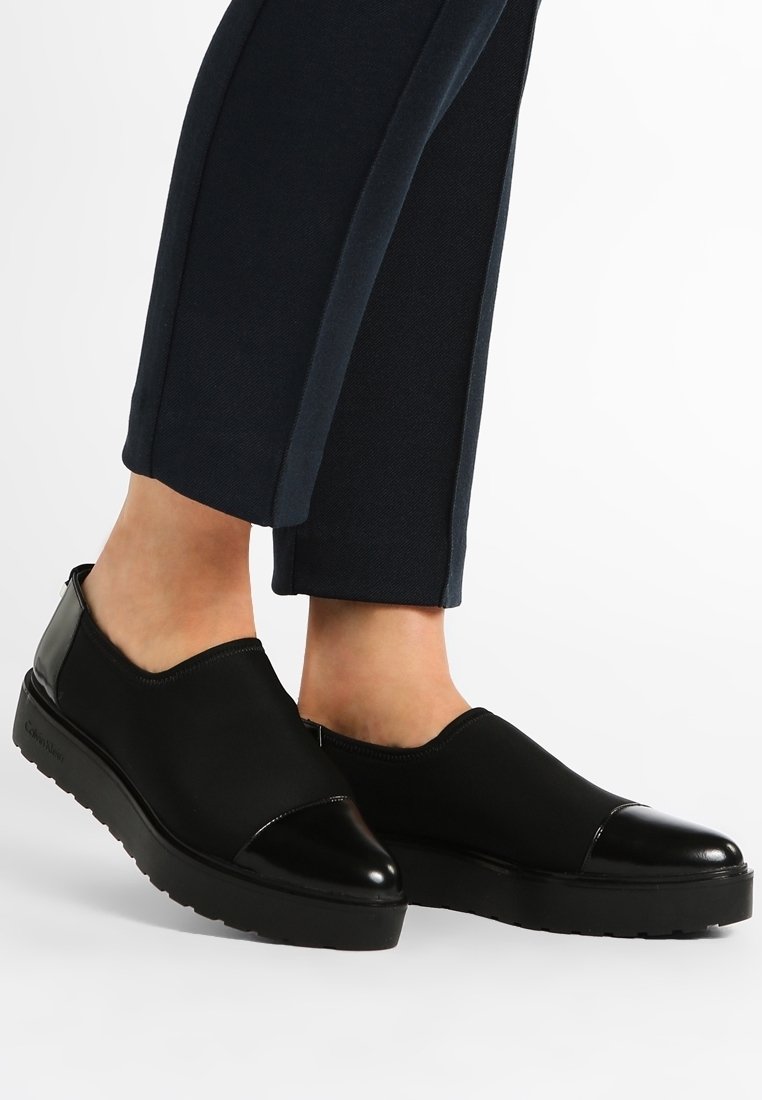 Zapatos negros de tipo slip-on fabricados con material sintético suave, con una puntera negra brillante y una suela texturada y flexible.