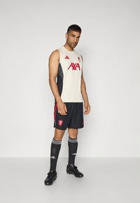 Sportuniform mit einem beigen ärmellosen Oberteil mit schwarzen Akzenten, rotem Logo und schwarzen Shorts mit roten Streifen; dazu schwarze Socken und Fußballschuhe.