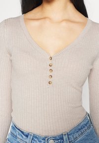 Nainen, jolla on päällään beige, ribbineuloksesta valmistettu V-kauluksinen pitkähihainen paita, jossa on kuusi nappia, yhdistettynä korkeavyötäröisiin sinisiin farkkuihin.