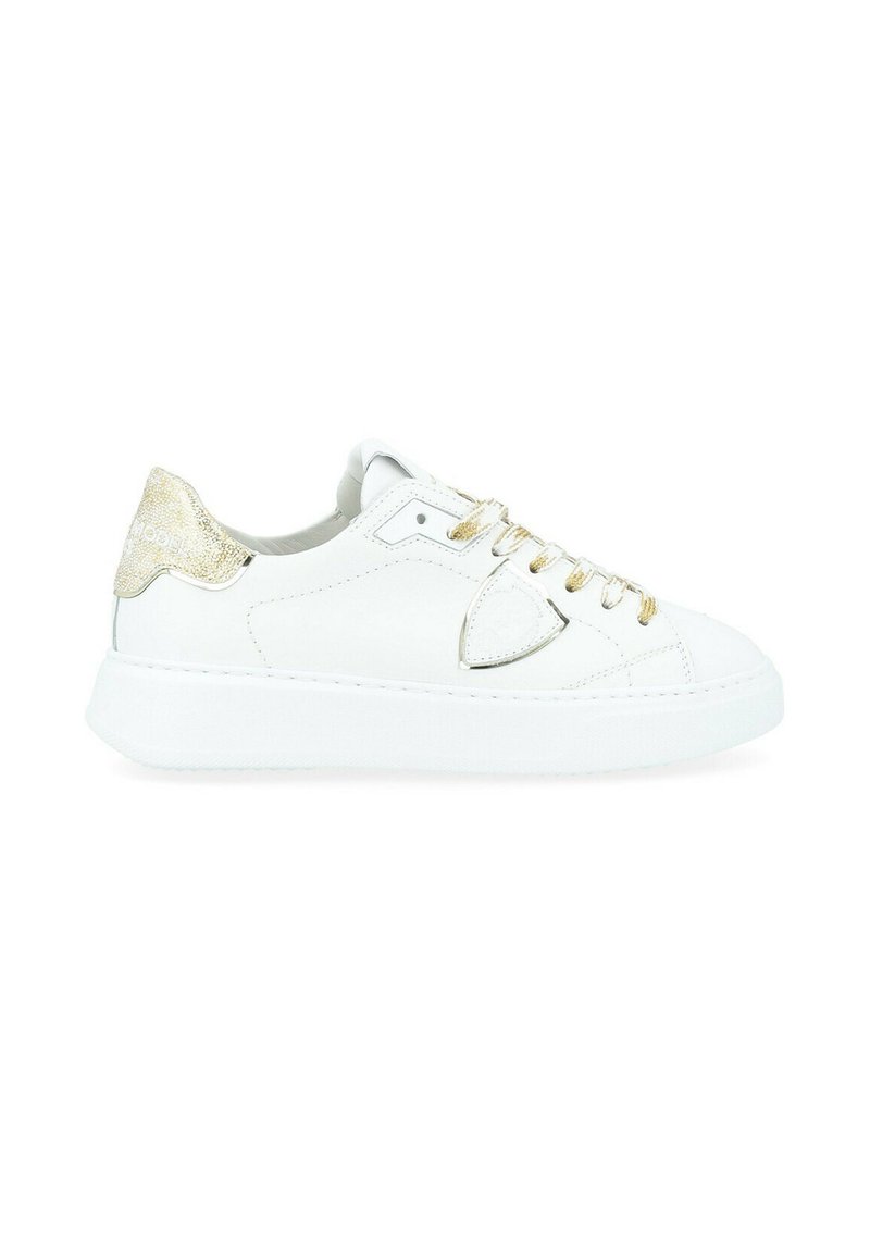 Baskets en cuir blanc avec un accent doré brillant au talon, semelle plate blanche et lacets dorés. Le panneau latéral présente une découpe triangulaire.