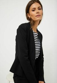 Černá bunda vyrobená z hladkého materiálu, s dobře padnoucím střihem a bez knoflíků. Nositá přes pruhovanou černo-bílou košili. Jednoduchý, moderní design.