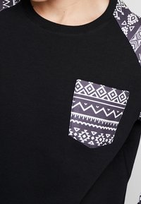 Felpa nera con maniche raglan decorate e una tasca. La tasca presenta disegni geometrici bianchi su uno sfondo scuro.