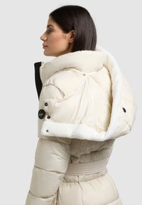 Gepolsterte, cremefarbene Jacke mit hohem Kragen, mit Fleece gefütterter Kapuze, Druckknöpfen und Tunnelzug in der Taille. Glatte, glänzende Textur.