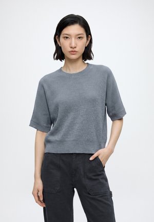 Donna con capelli corti neri che indossa un top grigio a maniche corte in maglia e pantaloni neri, in piedi con una mano in tasca su sfondo bianco.