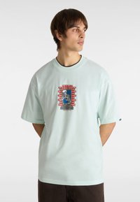 T-shirt mint verde a maniche corte con una stampa grafica di uno scheletro in una finestra di mattoni, accompagnata da un testo sotto l'immagine. Design con scollo rotondo.