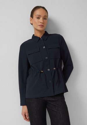 Femme portant une veste boutonnée bleu marine avec deux poches poitrine et une taille à cordon, associée à un jean foncé, debout contre un mur gris.