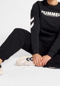 Sudadera negra con texto "HUMMEL", combinada con pantalones deportivos negros y zapatillas beige con rayas negras en ángulo. Atuendo deportivo cómodo.