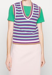 Gilet en tricot avec un col en V, présentant des rayures horizontales vertes, blanches, violettes et bleues, porté par-dessus un t-shirt vert, associé à une jupe rose pâle.
