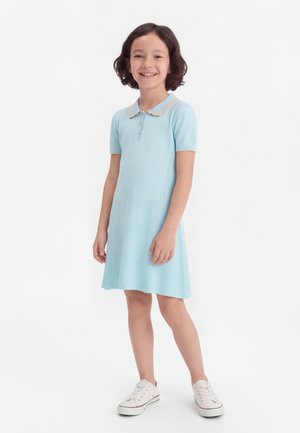 Κοπέλα με κοντά σκούρα μαλλιά, που φοράει ανοιχτό μπλε κοντομάνικο φόρεμα με γιακά και λευκά αθλητικά παπούτσια, χαμογελαστή, στέκεται σε λευκό φόντο.