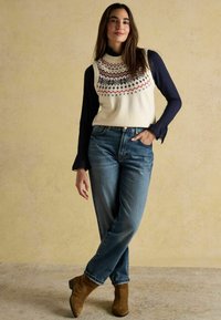 Ivory adīts veste ar daudzkrāsainiem ģeometriskiem rakstiem, kas valkāta virs tumši zila garām piedurknēm kreklam, apvienota ar zilajiem džinsiem un brūnām zamša potīšu zābakiem.