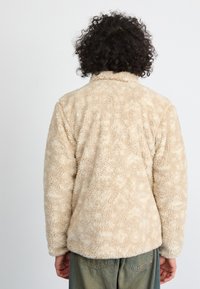 Person mit lockigem Haar, der eine beige Fleecejacke und grüne Hosen trägt, mit dem Rücken zu einem schlichten Hintergrund gewandt.