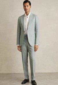 Costum din in albastru deschis cu rever tip notch, închidere cu un singur nasture, două buzunare frontale, purtat cu o cămașă albă cu nasturi și mocasini negri.
