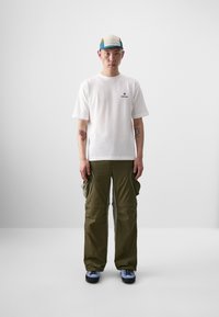 T-shirt blanche avec un petit logo, pantalon cargo vert olive avec des poches latérales, et chaussures bleues avec un capuchon à motifs.