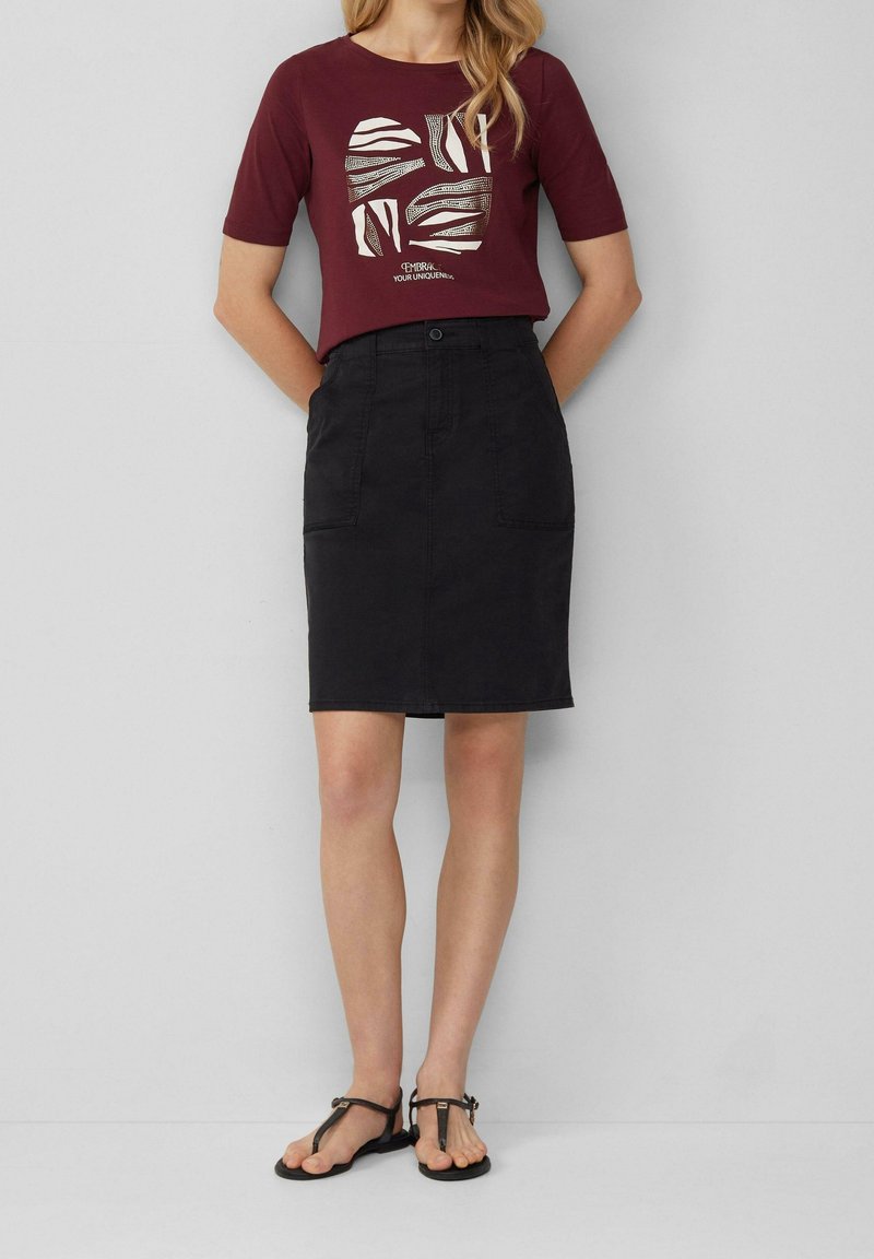 Femme portant un t-shirt graphique bordeaux, une jupe noire longueur genou avec poches avant, et des sandales plates noires, se tenant contre un mur gris uni.