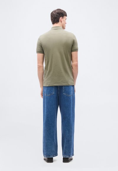 Homme de dos portant un polo vert olive, un jean bleu ample et des chaussures noires sur un fond clair uni.