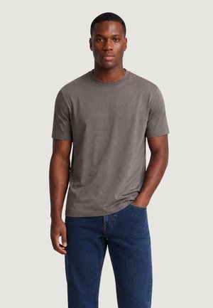 Mężczyzna ubrany w szarą krótką koszulkę Armani Exchange oraz niebieskie dżinsy, stoi z jedną ręką w kieszeni, zwrócony przodem.