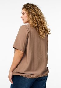 Short-sleeved, brown top made of lightweight fabric, featuring a rounded neckline and a loose fit, paired with blue denim jeans.