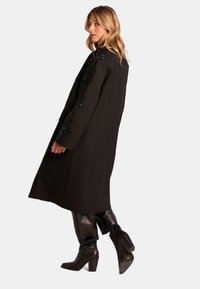 Abrigo negro largo con detalles bordados en las mangas, combinado con pantalones de cuero negros y botas de tacón. Textura suave y diseño a medida.