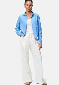 Chemise bleu clair à manches longues avec une poche sur la poitrine, débardeur blanc en dessous et pantalon en lin blanc ample, associés à des sandales marron.