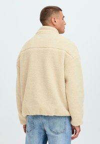 Chaqueta de forro polar color crema con cuello alto, superficie texturizada y corte relajado. Presenta mangas largas y un dobladillo recto. Combinada con jeans de color azul claro.