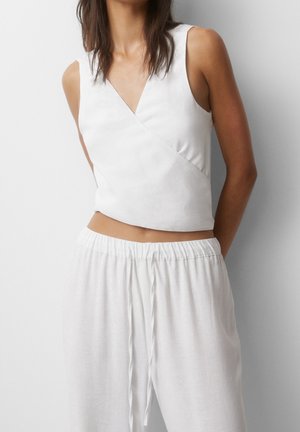 Kvinde iført hvid ærmeløs crop top med omslag, kombineret med løstsiddende hvide bukser med snøre, mod en ensfarvet baggrund.