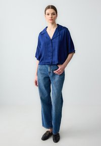 Blaue kurzärmlige Hemd mit Knopfleiste aus strukturiertem Stoff, kombiniert mit hochgeschnittenen blauen Jeans und schwarzen Slippern.