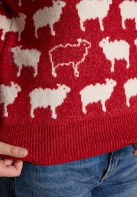 Roter Strickpullover mit weißen Schafmustern, ein umrandetes Schaf, getragen zu blauen Jeans, Hände halten den Saum des Pullovers.
