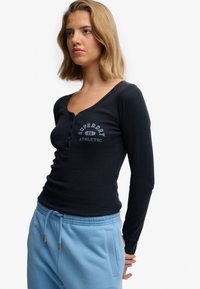 Haut noir à manches longues avec un col en V profond, une patte de boutonnage, et un texte imprimé gris indiquant "Superdry 86 Athletic" sur la poitrine.