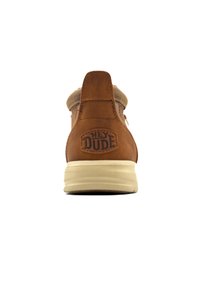 Botte de cheville en suede marron avec un col beige, semelle extérieure crème plate et logo "HEY DUDE" à l'arrière. Texture lisse avec des coutures minimales.