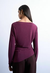 Femme aux longs cheveux foncés, portant un haut violet à manches longues et un pantalon foncé, debout de dos sur un fond clair uni.