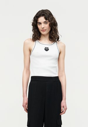 Γυναίκα με καστανά σγουρά μαλλιά που φοράει λευκό αμάνικο τοπ με μαύρα γαζιά και έμβλημα, συνδυασμένο με ψηλόμεσο μαύρο παντελόνι.