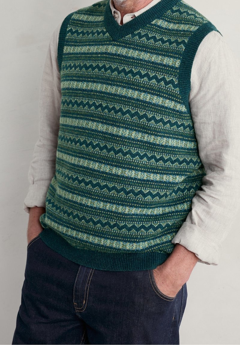 Homme portant un gilet tricoté vert à motifs par-dessus une chemise blanche à manches longues retroussées et un jean bleu foncé, les mains dans les poches.