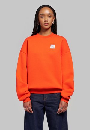 Mujer con pelo largo trenzado, vistiendo una sudadera naranja con un logo blanco y vaqueros azul oscuro, de pie frente a un fondo gris liso.