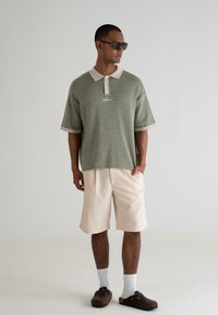 Green textured polo shirt with a light collar, beige shorts, and brown clogs, paired with white socks and sunglasses. Casual outfit displayed.
