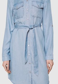 Ljusblå denimklänning med långa ärmar, knäppdetaljer, två bröstfickor och ett knytbart midjeband. Slät textur och rundad fåll.