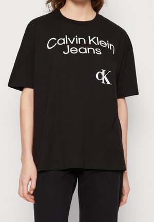 Personne portant un t-shirt noir Calvin Klein Jeans avec un logo blanc et un pantalon noir, debout devant un fond uni.