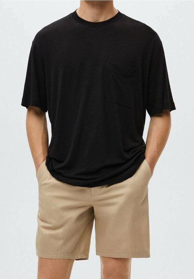 Mand iført en løs sort T-shirt med brystlomme og beige shorts, hænderne i lommerne, stående foran en ensfarvet lys baggrund.