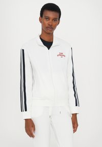 Witte zip-up jas met marineblauwe strepen op de mouwen, voorzien van rode geborduurde tekst "MILANO GYMNASIUM", gemaakt van een zachte stof.